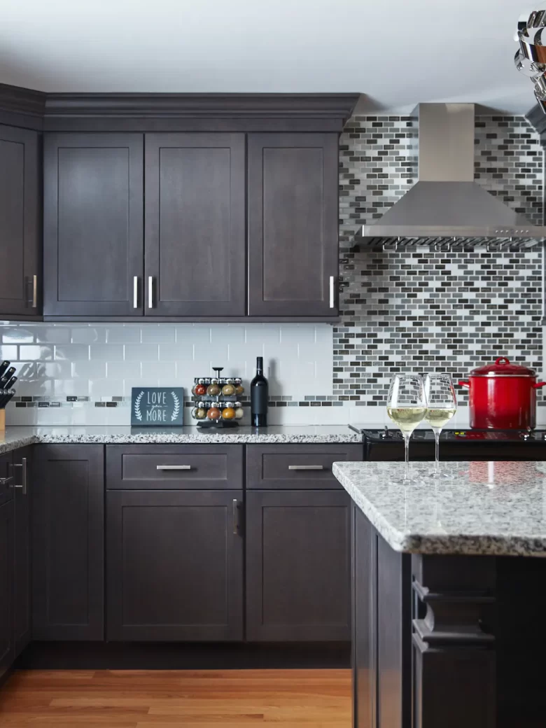 Modern kitchen with dark cabinets, mosaic backsplash, red pot, and two glasses of white wine on the counter.