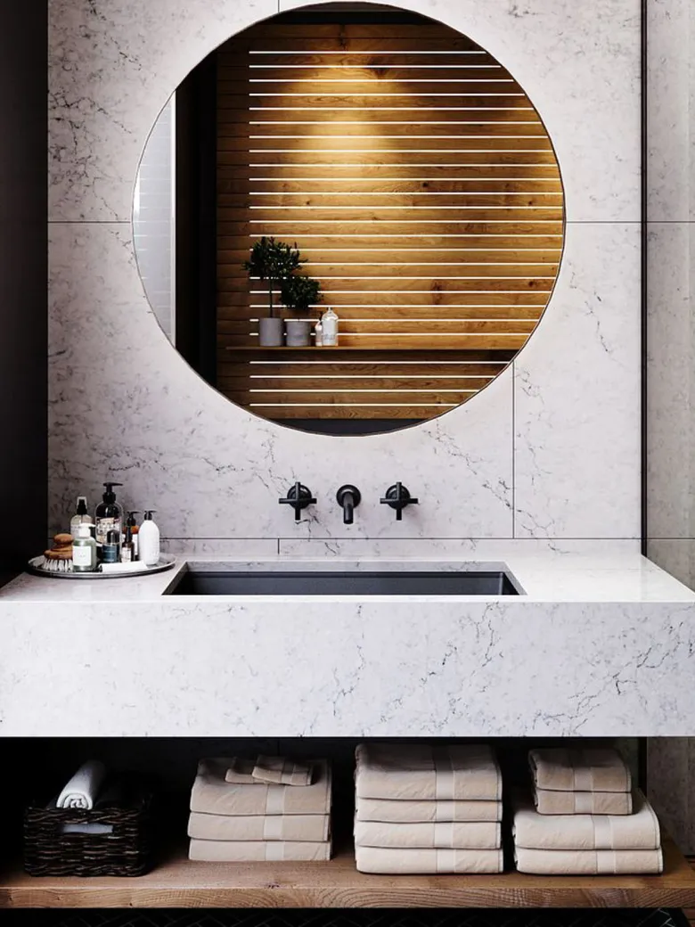 Modern bathroom with a marble sink, round mirror, toiletries, and folded towels on shelves below.