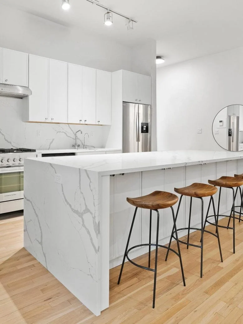 Modern white kitchen with marble island, wood floors, three wooden stools, and stainless steel appliances.