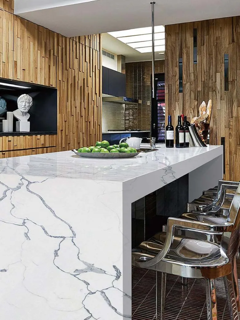 Modern kitchen with a marble island, metal chairs, wood panel walls, and a bowl of green apples.