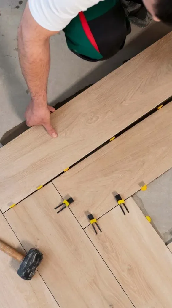 A person installs wooden floor panels, using spacers and a rubber mallet for alignment.